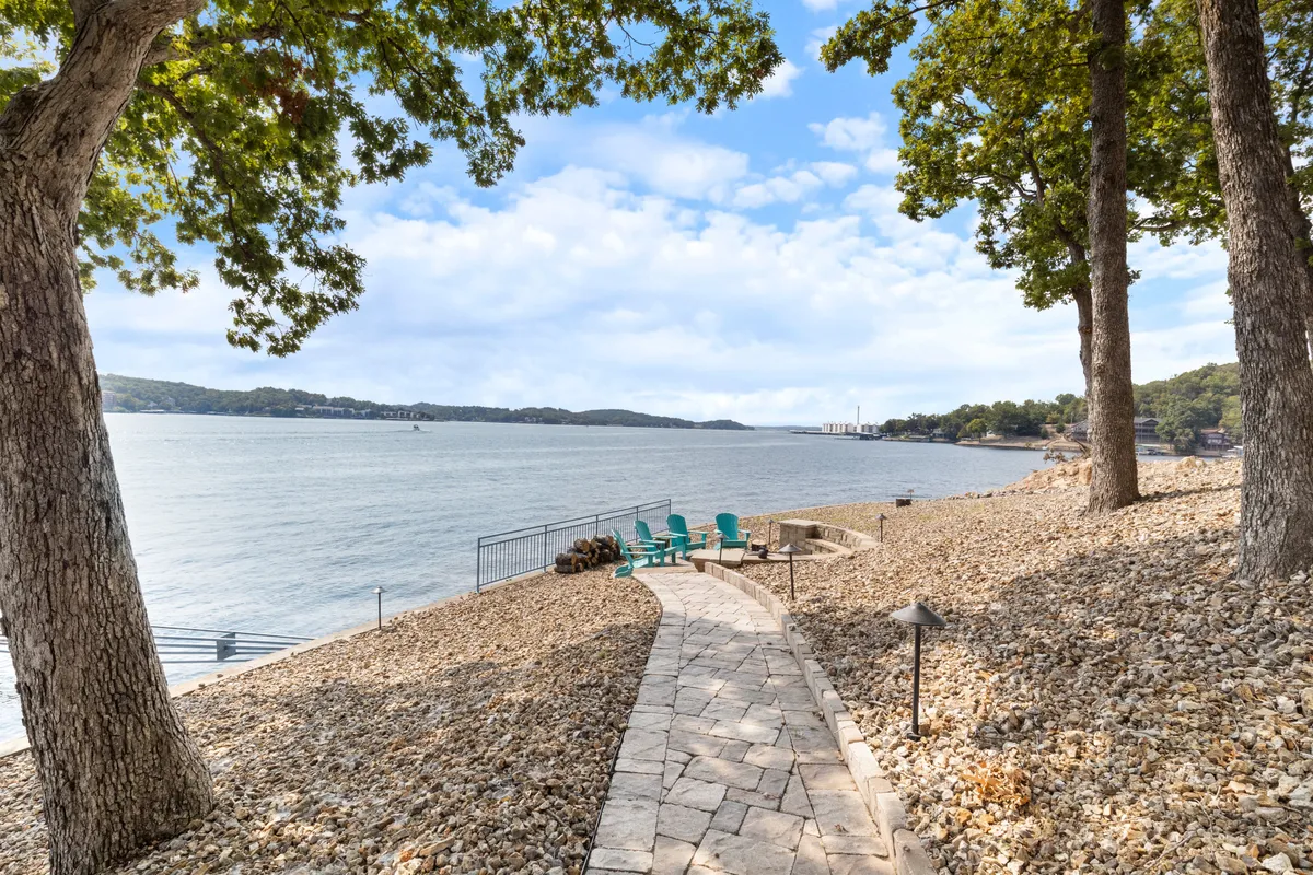 A scenic pathway lined with trees leads to a set of Adirondack chairs positioned at the water’s edge. The scene emphasizes the connection between the property and the outdoors, with ample room to walk, play, or sit together while enjoying lake breezes and natural beauty.