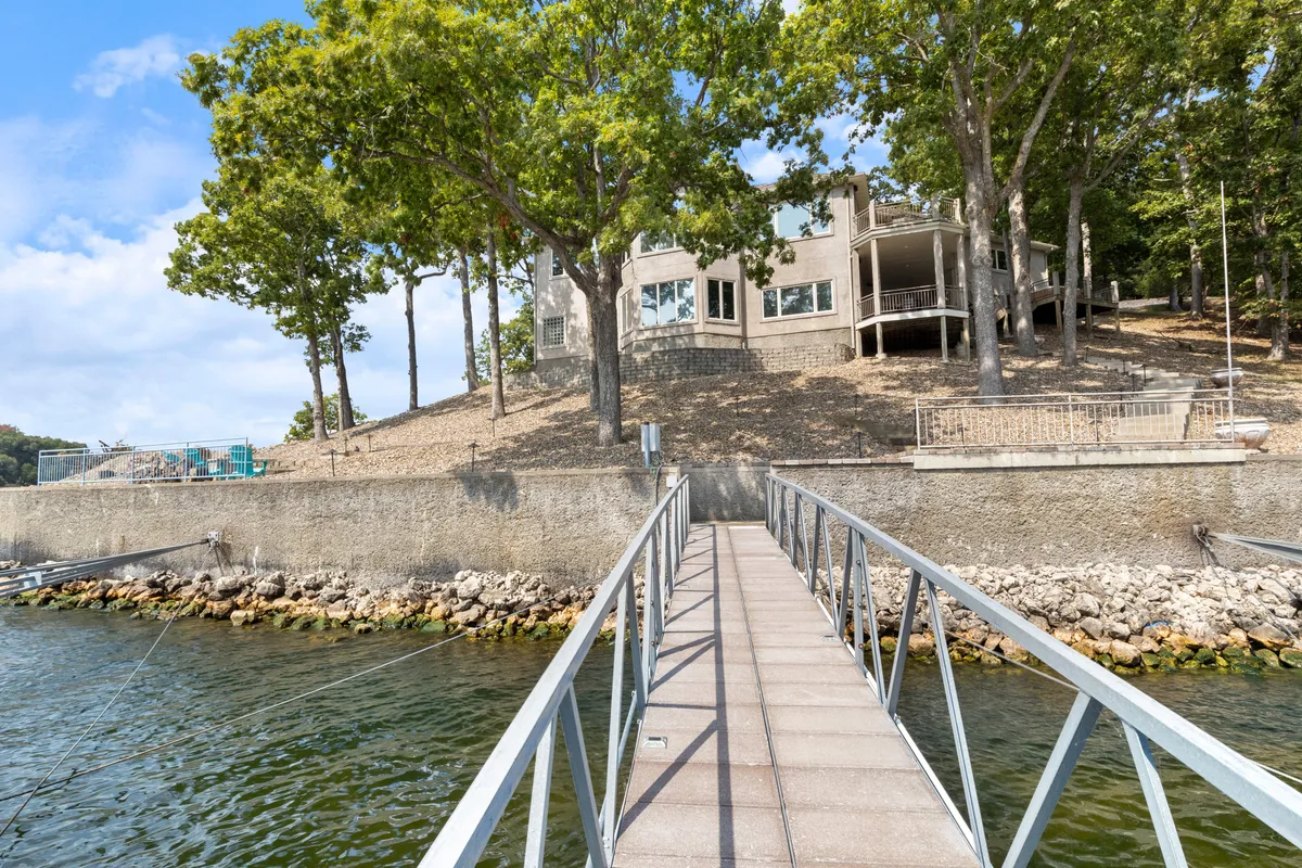 This image captures a dock stretching out into the lake, with the house visible on a hillside and trees framing the water view. The tranquil atmosphere hints at activities like fishing, boating, and swimming, while the sunset light suggests a picturesque end to the day.