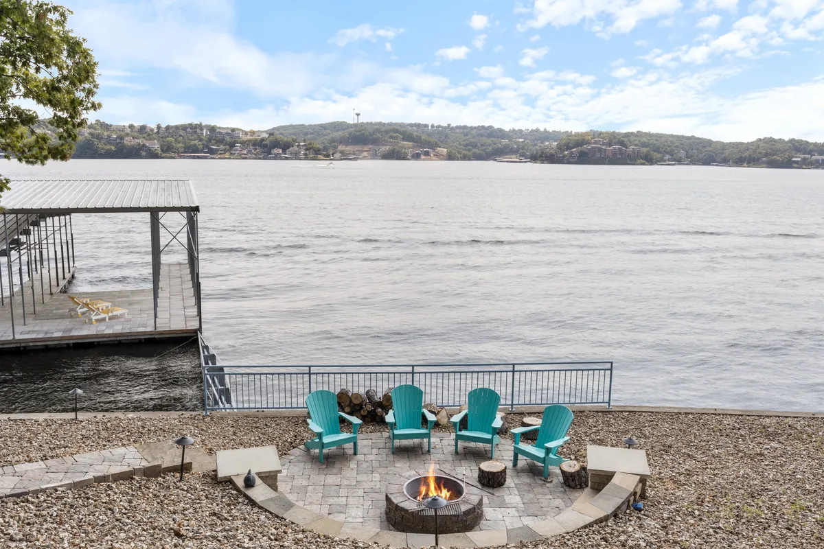 This image features a lakeside fire pit surrounded by Adirondack chairs, with a sweeping view of the lake and dock in the background. The inviting seating arrangement is perfect for group gatherings, storytelling, and evening relaxation as the sun sets over the water.
