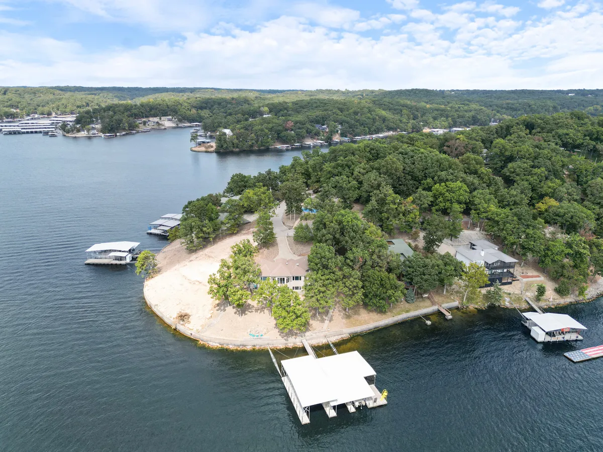 A wider shot of the lakefront property showing the main structure nestled among greenery, a spacious beach area, multiple docks extending into the lake, and people enjoying swimming, boating, and picnicking near the shore.