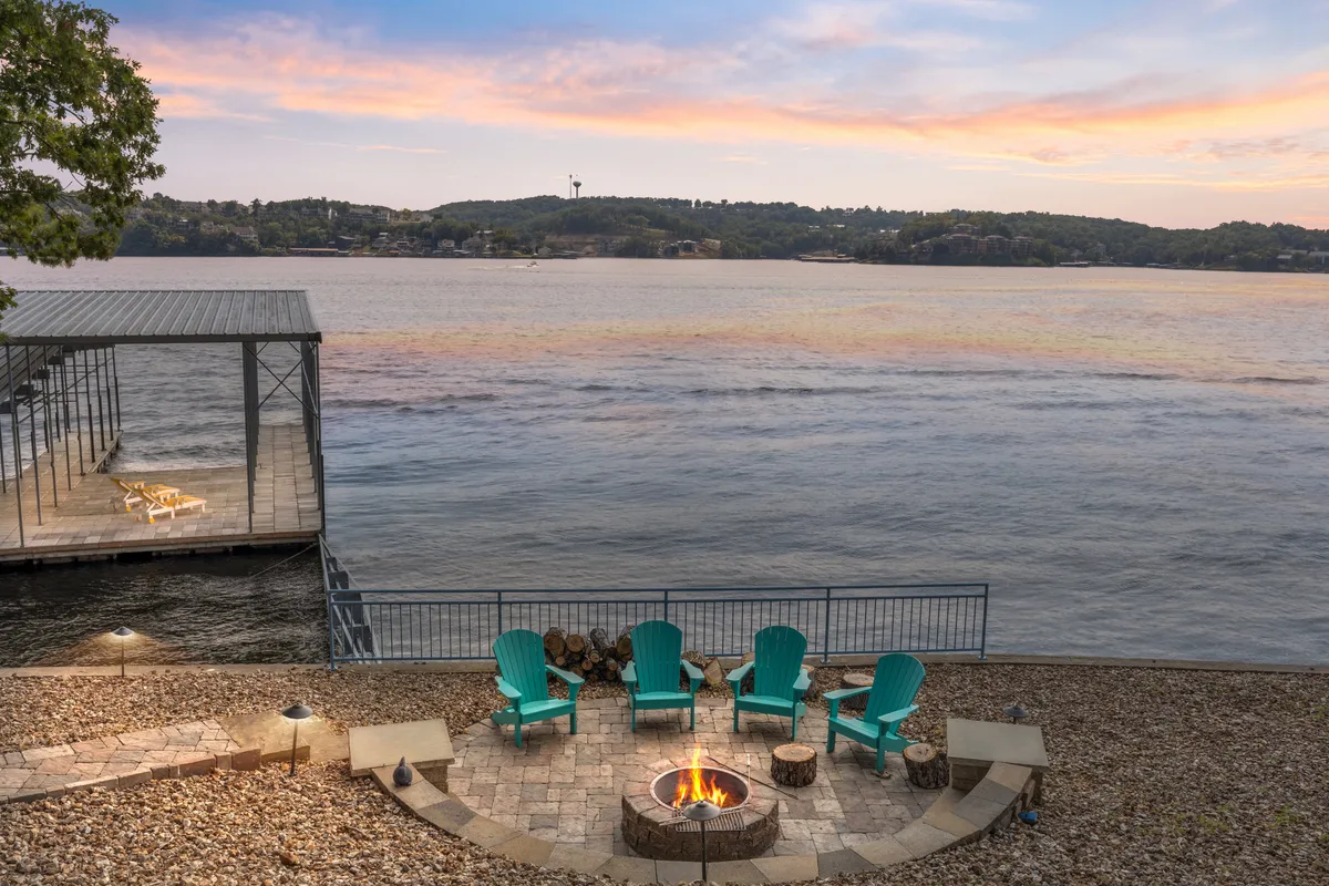 The image showcases the home's scenic lakefront setting at sunset, with a cozy fire pit surrounded by Adirondack chairs, a private dock extending over the shimmering water, and tranquil views that invite relaxation. The inviting glow of the fire and the expansive lake horizon set a serene, welcoming tone.