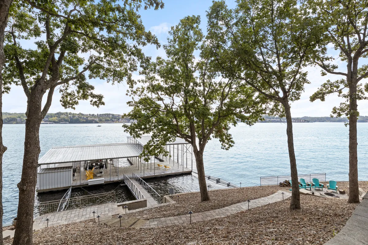 A close-up exterior shot of the dock area with patio seating, trees for shade, and clear views of the serene lake. The dock is well-suited for launching boats and relaxing by the water, with ample space for gathering with friends or fishing off the edge.
