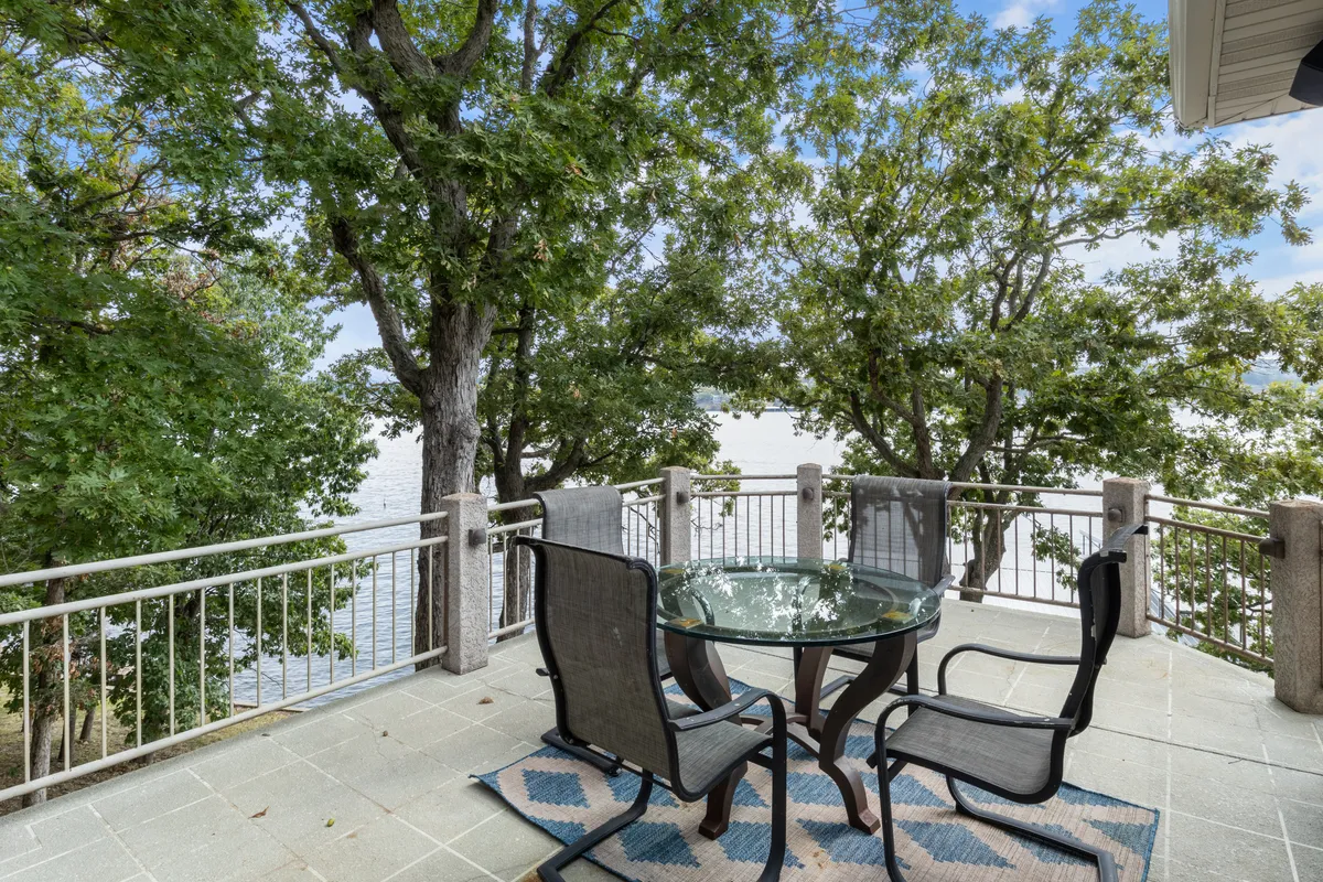 This image features a glass-topped table with outdoor chairs set on a deck, surrounded by lush greenery and overlooking the lake. The peaceful ambiance is perfect for morning coffee, reading, or simply enjoying nature.